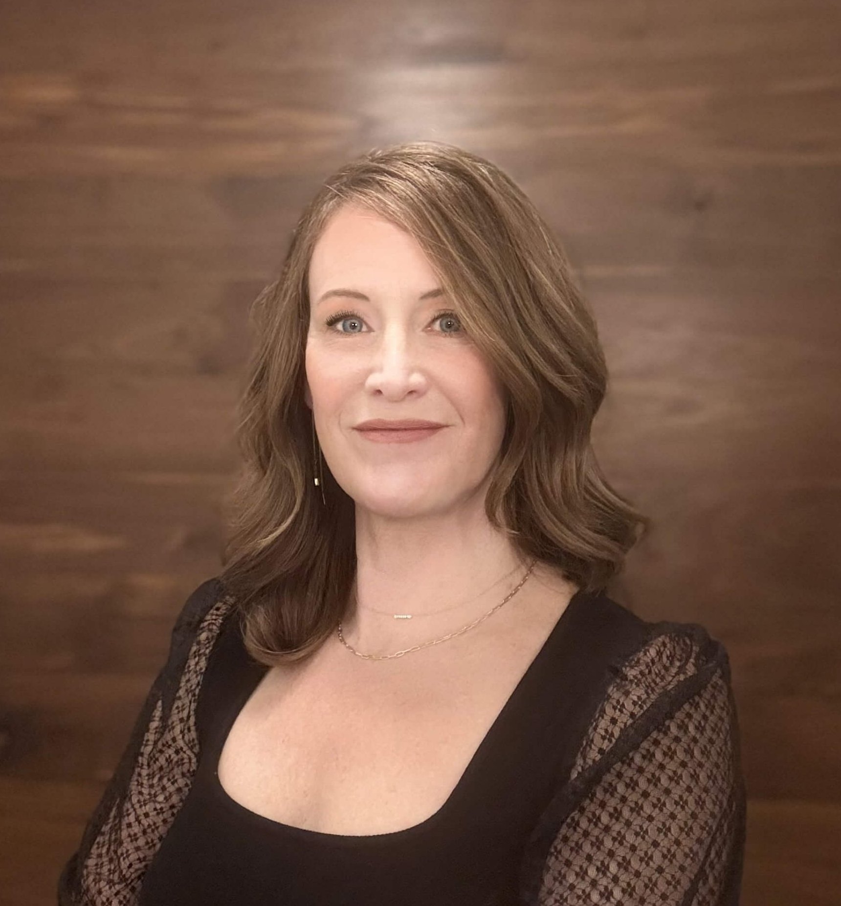 Professional headshot of a tattoo artist in front of a warm wood background, with soft wavy hair and a black top.